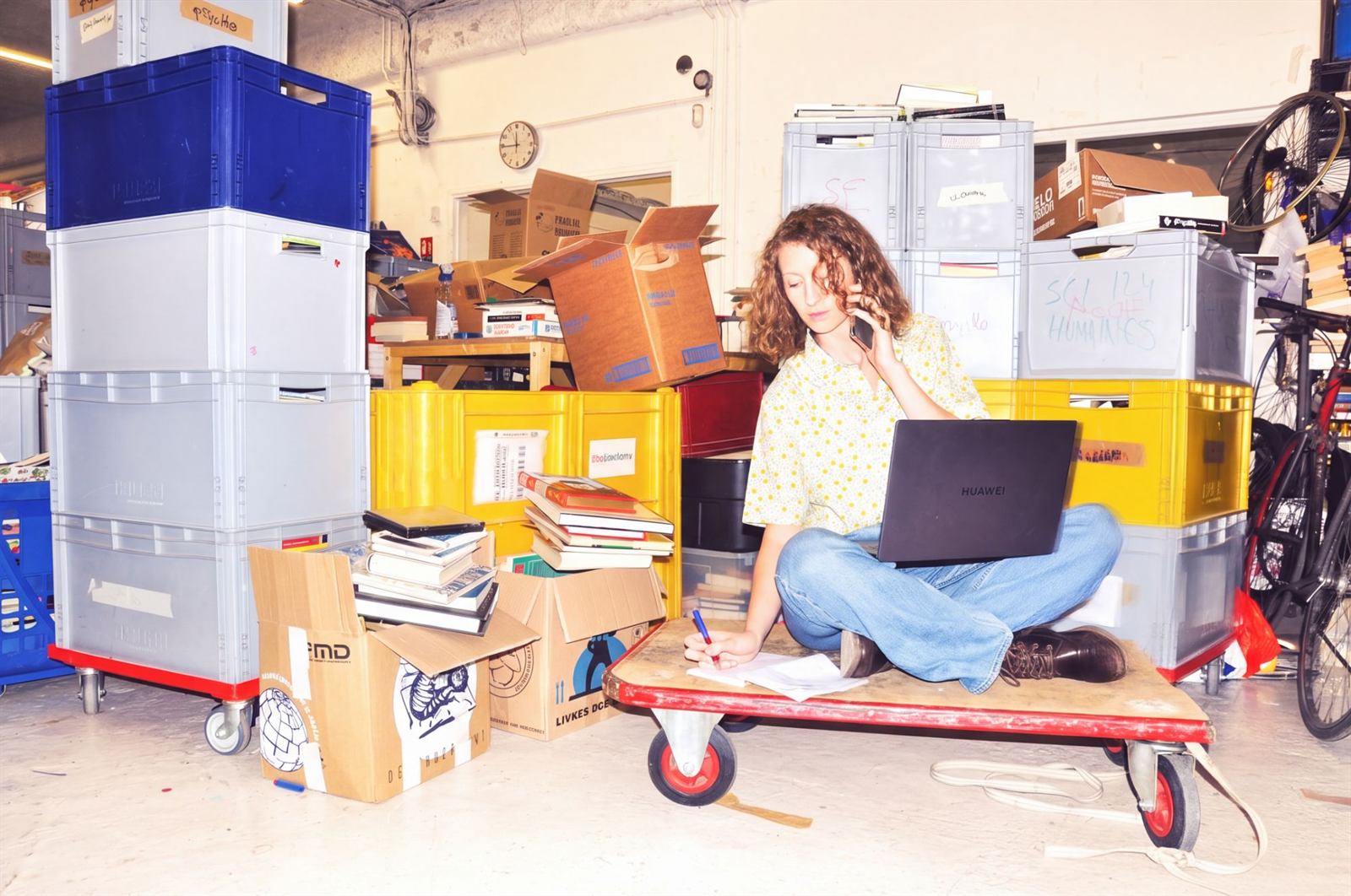 Caroline Grosjean au téléphone, assise en tailleur sur un chariot, entourée de caisses en ressourcerie